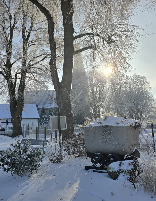 Verschneite Bergbaulore mit Raureif vor der Adenstedter Kirche im winterlichen Gegenlicht